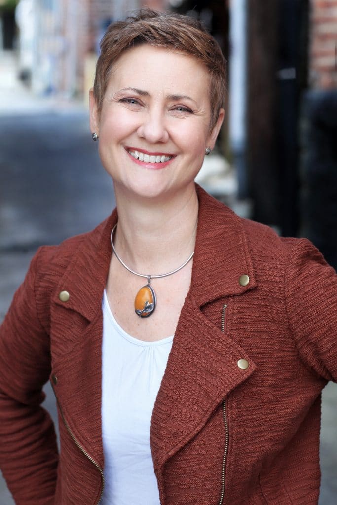 Angie Flynn-McIver smiles at the camera. She wears a dark red-orange jacket, white scoop neck shirt and orange stone necklace.